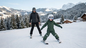 Smiling child in a ski suit snowploughing down a beginner slope, with a parent skiing behind