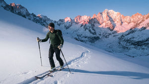 Ski tourer in a green merino wool base layer skinning up a snowy slope at sunrise, pink peaks in the background